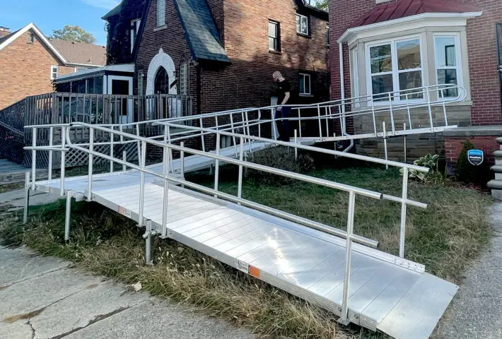 A metal wheelchair ramp with handrails leads up to the entrance of a brick house, providing accessible entry from the sidewalk across a grassy front yard.