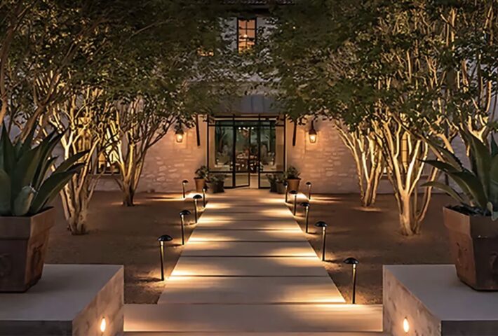 A well-lit stone pathway lined with small lights leads to a house entrance, flanked by potted agave plants and trees with warm outdoor lighting, creating an inviting nighttime atmosphere.