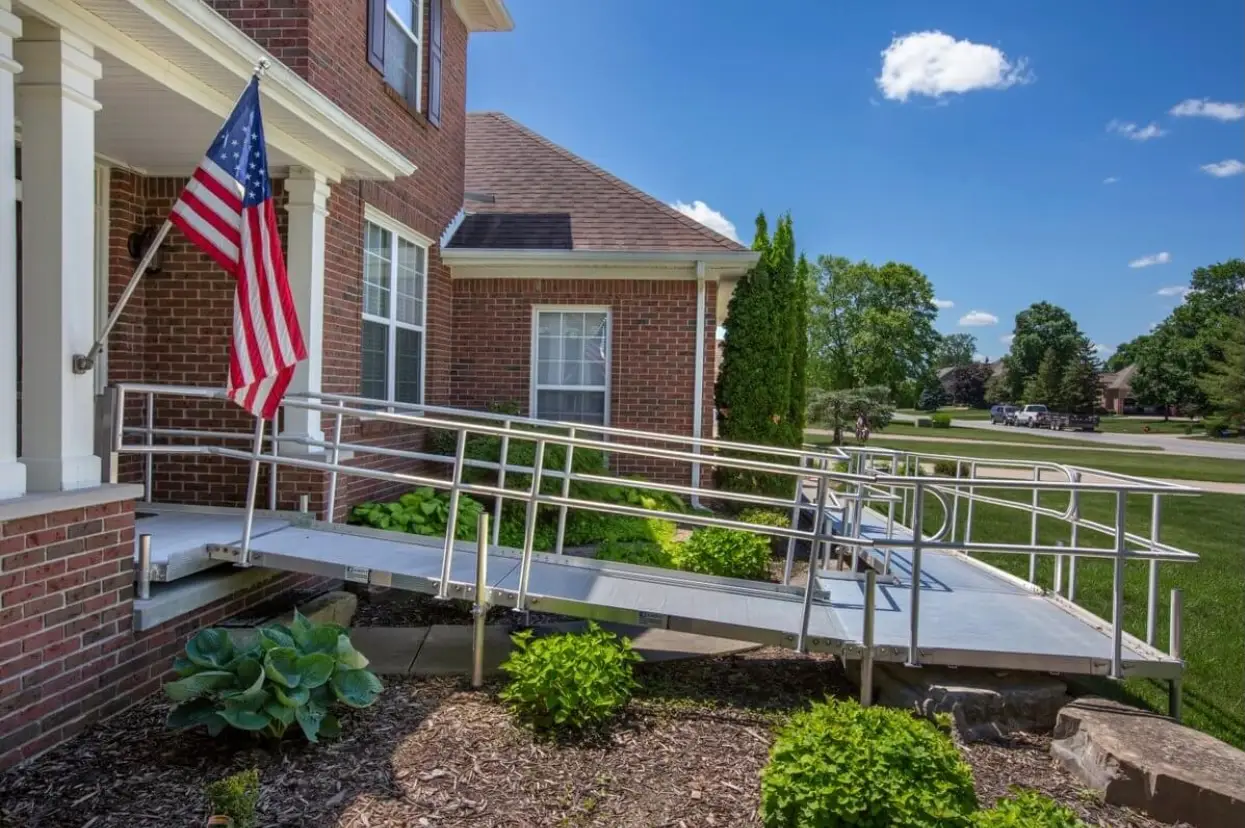 A silver metal wheelchair ramp leads to the entrance of a brick house. An American flag hangs by the door, with shrubs and green plants along the front yard under a sunny, blue sky.
