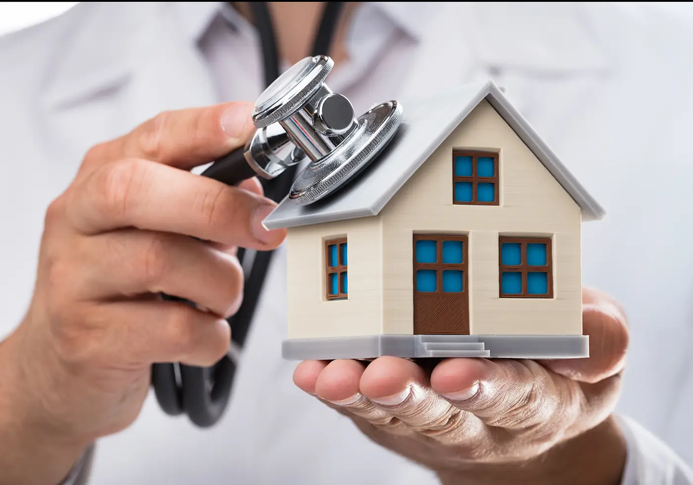 A person in a white coat uses a stethoscope to listen to a small model house, symbolizing a home health check or house inspection.