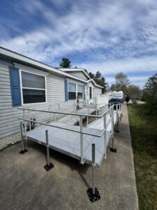 A modular aluminum wheelchair ramp is installed next to a sidewalk leading to the entrance of a single-story house with blue shutters. Trees and a van are visible in the background under a partly cloudy sky.