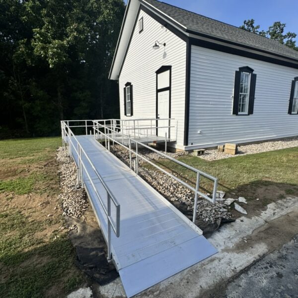 A metal wheelchair ramp with handrails leads to the entrance of a white building with black shutters, surrounded by grass and trees on a sunny day in Tyrone Township Michigan.