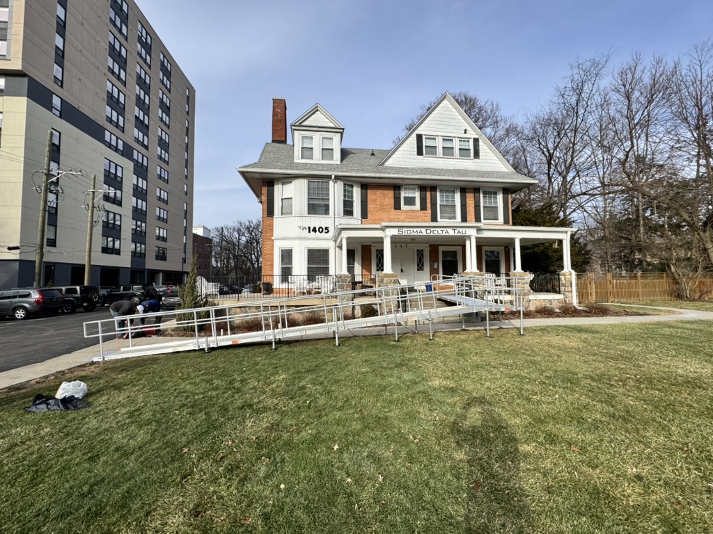 A large, two-story brick and gray house with a front porch and a long wheelchair ramp leading to the entrance. The house has a sign reading Sigma Delta Tau and the number 1405. There are trees and a grassy lawn in front on the campus of University of Michigan, Ann Arbor.