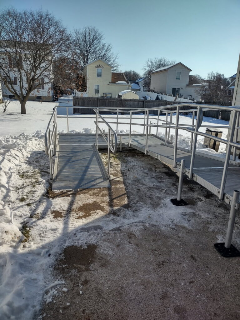 A metal wheelchair ramp leads down to a snowy backyard with patches of cleared ground. Several houses, leafless trees, and a wooden fence are visible in the background under a clear blue sky in Grand Rapids Michigan.