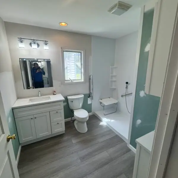Accessible bathroom with light wood flooring, white vanity, toilet, and a walk-in shower with a shower chair and grab bar. A person is visible in the mirror, and natural light comes from a window above the toilet, installed in Grand Blanc Michigan.