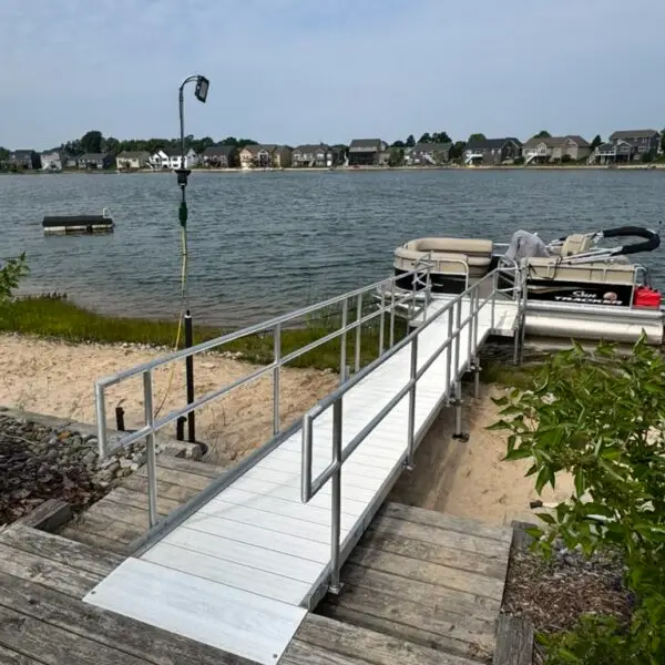 A metal ramp with railings leads from wooden steps down to a sandy lakeshore with a pontoon boat docked at the end; houses and a floating platform are visible across the lake under a partly cloudy sky in Hudsonville Michigan.