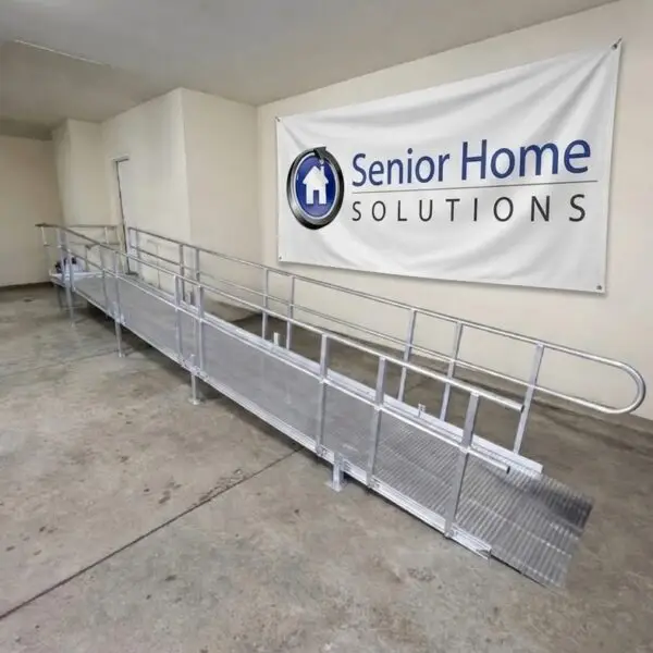 A long metal wheelchair ramp with railings is installed indoors next to a wall in Milford Michigan. A large banner on the wall reads Senior Home Solutions with a house logo. The floor is concrete.