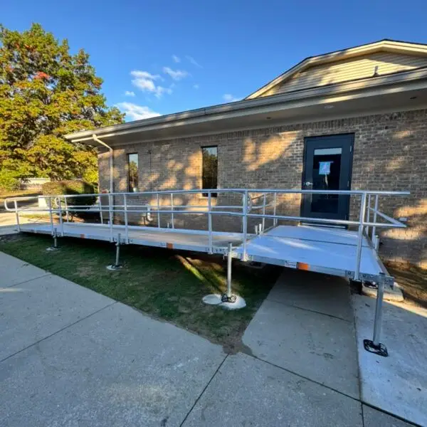 A metal wheelchair ramp with handrails leads up to the entrance of a brick building under a clear blue sky in Waterford Michigan. The ramp crosses over a grassy patch and connects to the sidewalk.
