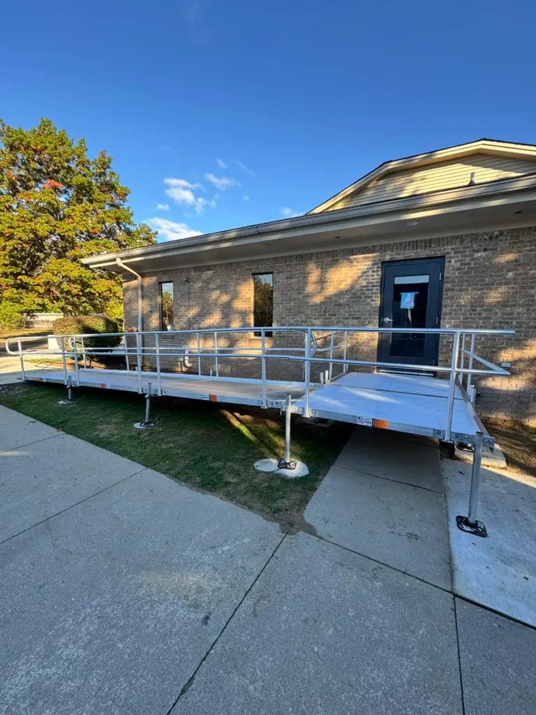 A metal wheelchair ramp with handrails leads up to the entrance of a brick building under a clear blue sky in Waterford Michigan. The ramp crosses over a grassy patch and connects to the sidewalk.
