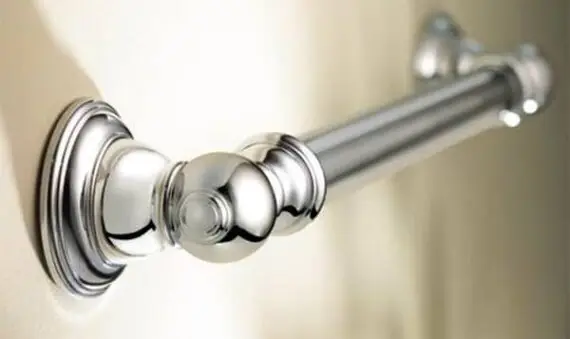 A close-up view of a shiny, silver metal grab bar mounted horizontally on a beige wall, often used for support in bathrooms or showers.