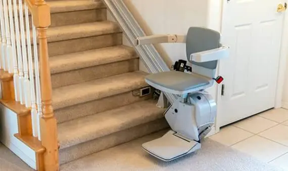 A motorized stair lift with a gray seat and armrests is installed along a carpeted staircase, allowing for easy access between floors. The lift is positioned at the base of the stairs near a white door.