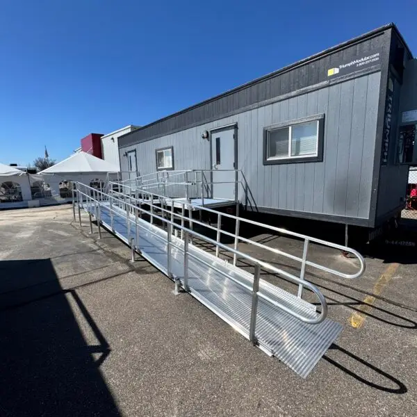 A gray portable building with steps and a long metal wheelchair-accessible ramp leading to its entrance, situated in a sunny parking lot in Birch Run Michigan.