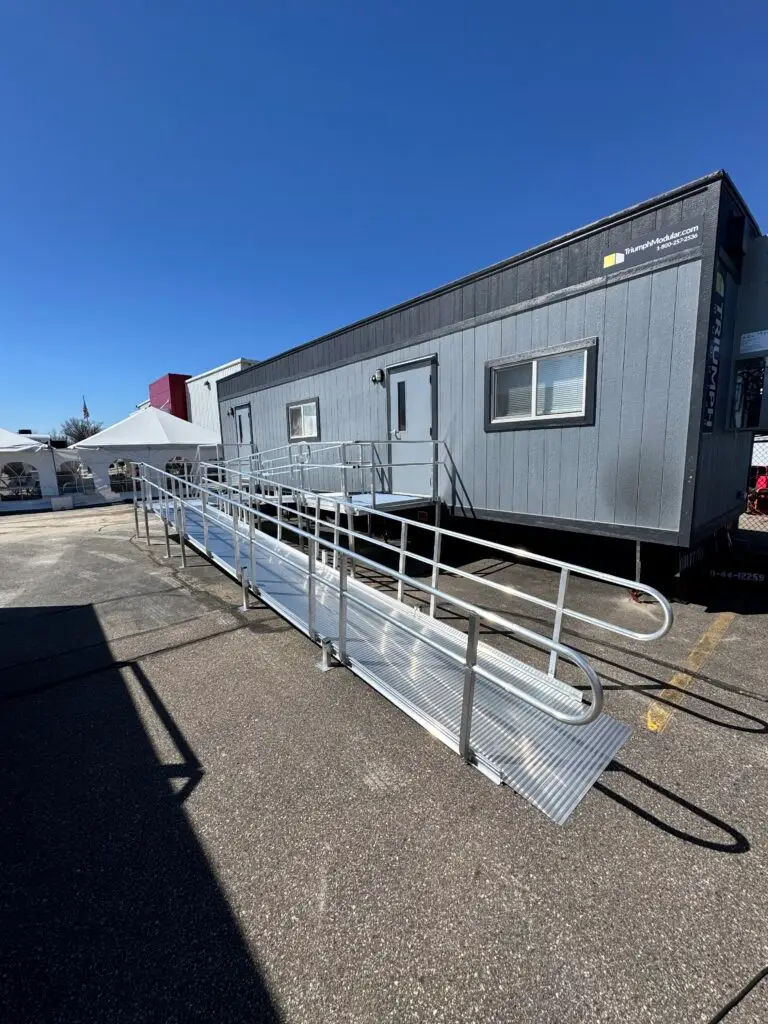 A gray portable building with steps and a long metal wheelchair-accessible ramp leading to its entrance, situated in a sunny parking lot in Birch Run Michigan.