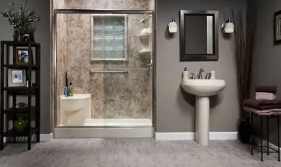 A modern bathroom with a walk-in shower, glass doors, and beige tile, a white pedestal sink with a mirror above it, wall-mounted lights, and shelves with decor and towels on a carpeted floor.