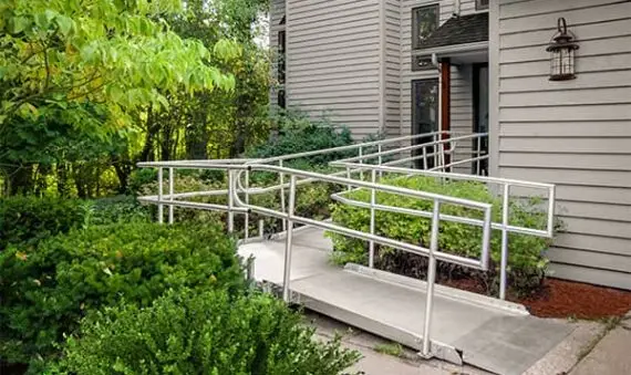 A concrete wheelchair ramp with metal railings leads to the entrance of a gray building, surrounded by bushes and greenery.