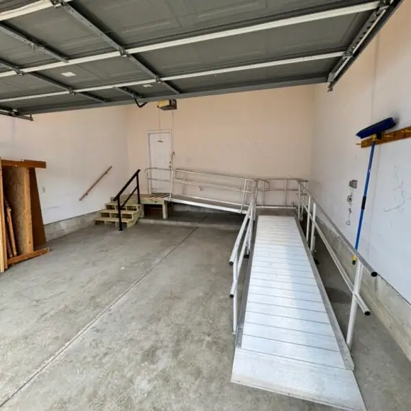 This Ann Arbor wheelchair ramp installation is in a clean garage with an open door contains a metal wheelchair ramp and stairs leading to a white interior door. The walls are mostly bare, with some shelves and a broom on the right side.