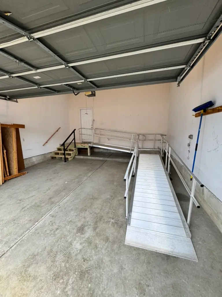 This Ann Arbor wheelchair ramp installation is in a clean garage with an open door contains a metal wheelchair ramp and stairs leading to a white interior door. The walls are mostly bare, with some shelves and a broom on the right side.