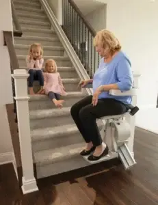 An older woman uses a stair lift on a carpeted staircase while two young girls sit and smile on the steps beside her. The setting appears to be a cozy home interior.