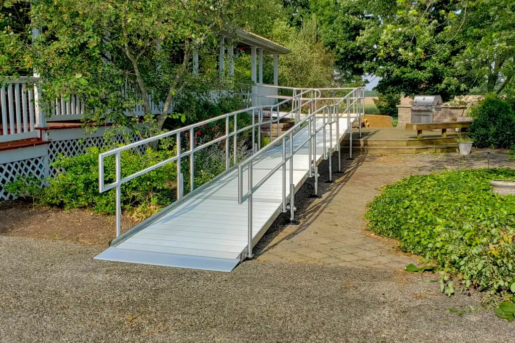 A metal wheelchair ramp with handrails leads from a paved path up to the porch of a house surrounded by greenery and trees.