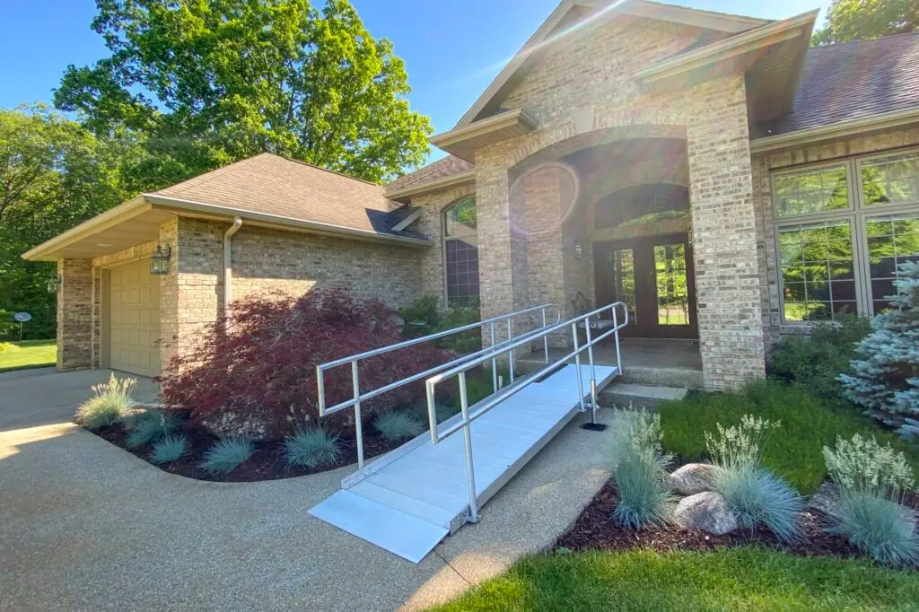 A brick house with large windows and a landscaped garden features a silver wheelchair ramp leading from the driveway to the front door under a covered porch.