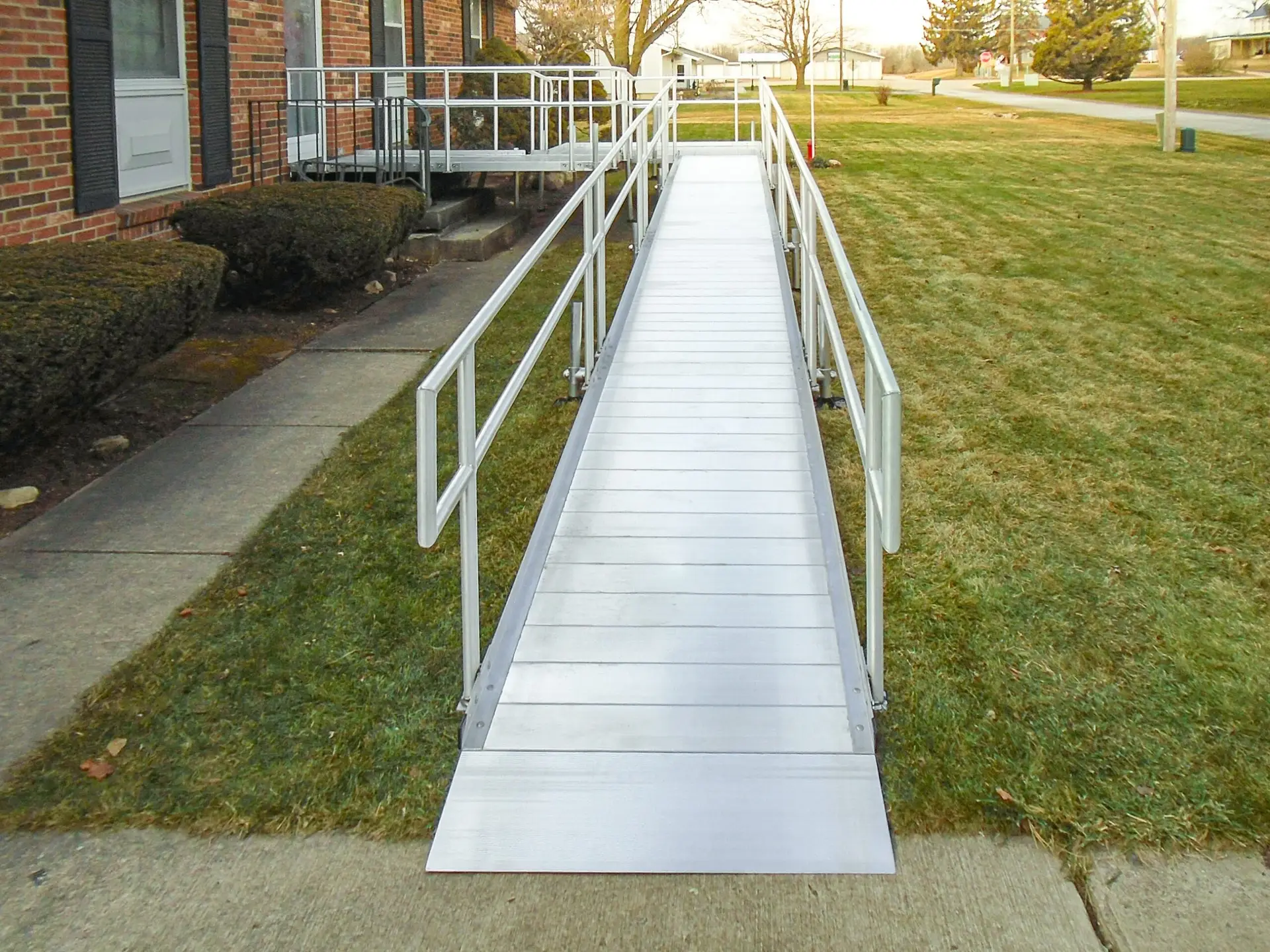 A metal wheelchair ramp with handrails on both sides leads from a sidewalk up to the entrance of a brick building, situated next to a grassy lawn.