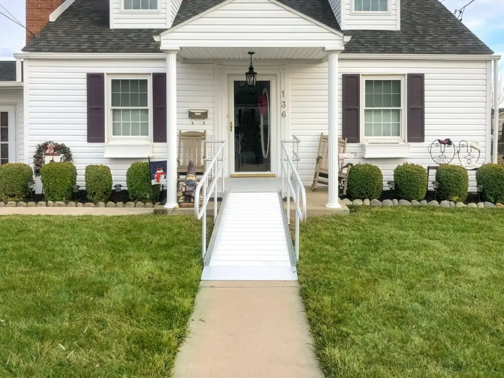 A white house with a front porch, purple shutters, and a ramp leading up to the front door. The yard is green and neatly trimmed, and there are bushes and small decorations near the entrance.