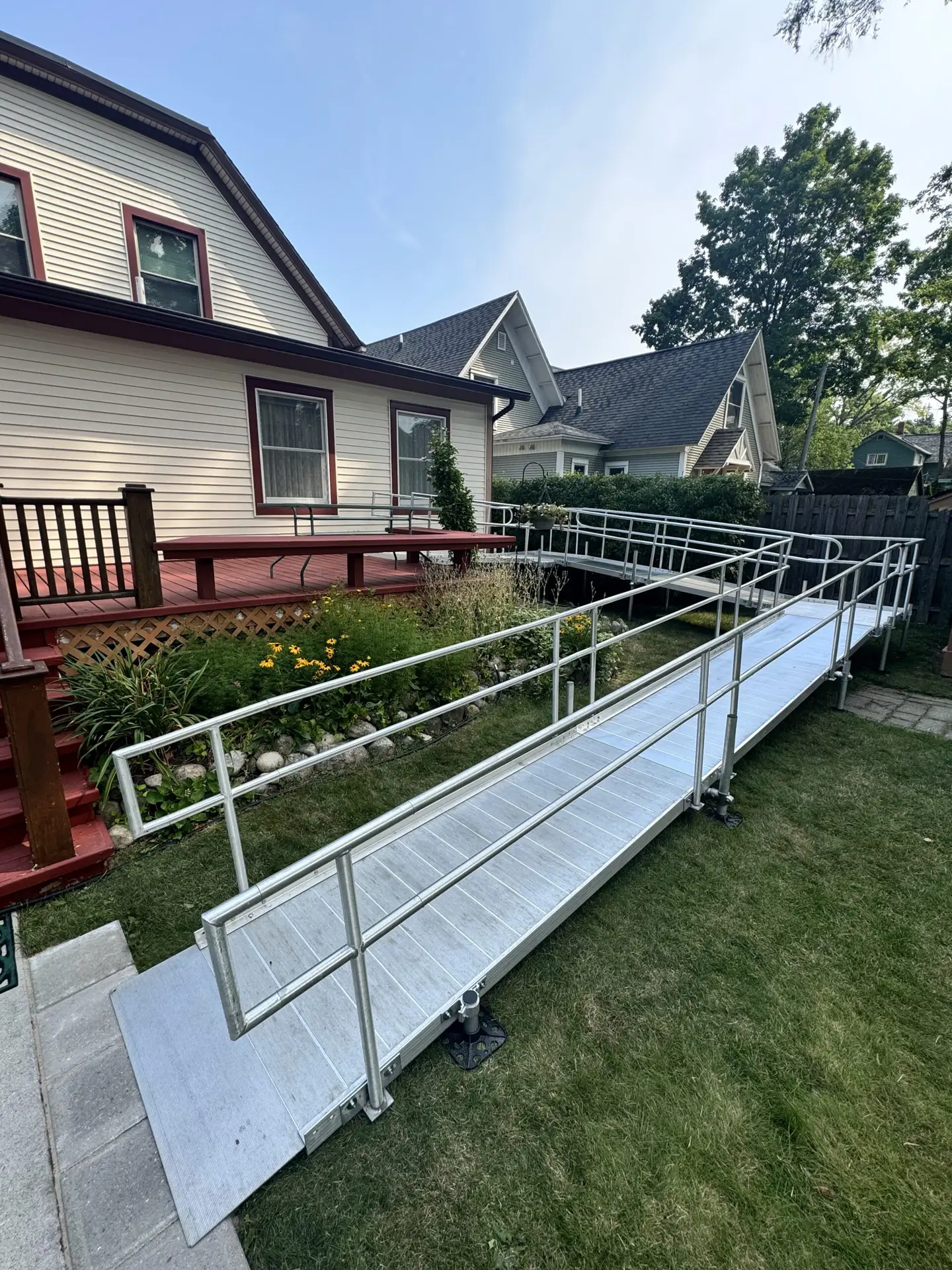 A long, silver metal wheelchair ramp with railings leads from a grassy yard up to the porch of a beige house with red trim, surrounded by plants and neighboring homes in the background.
