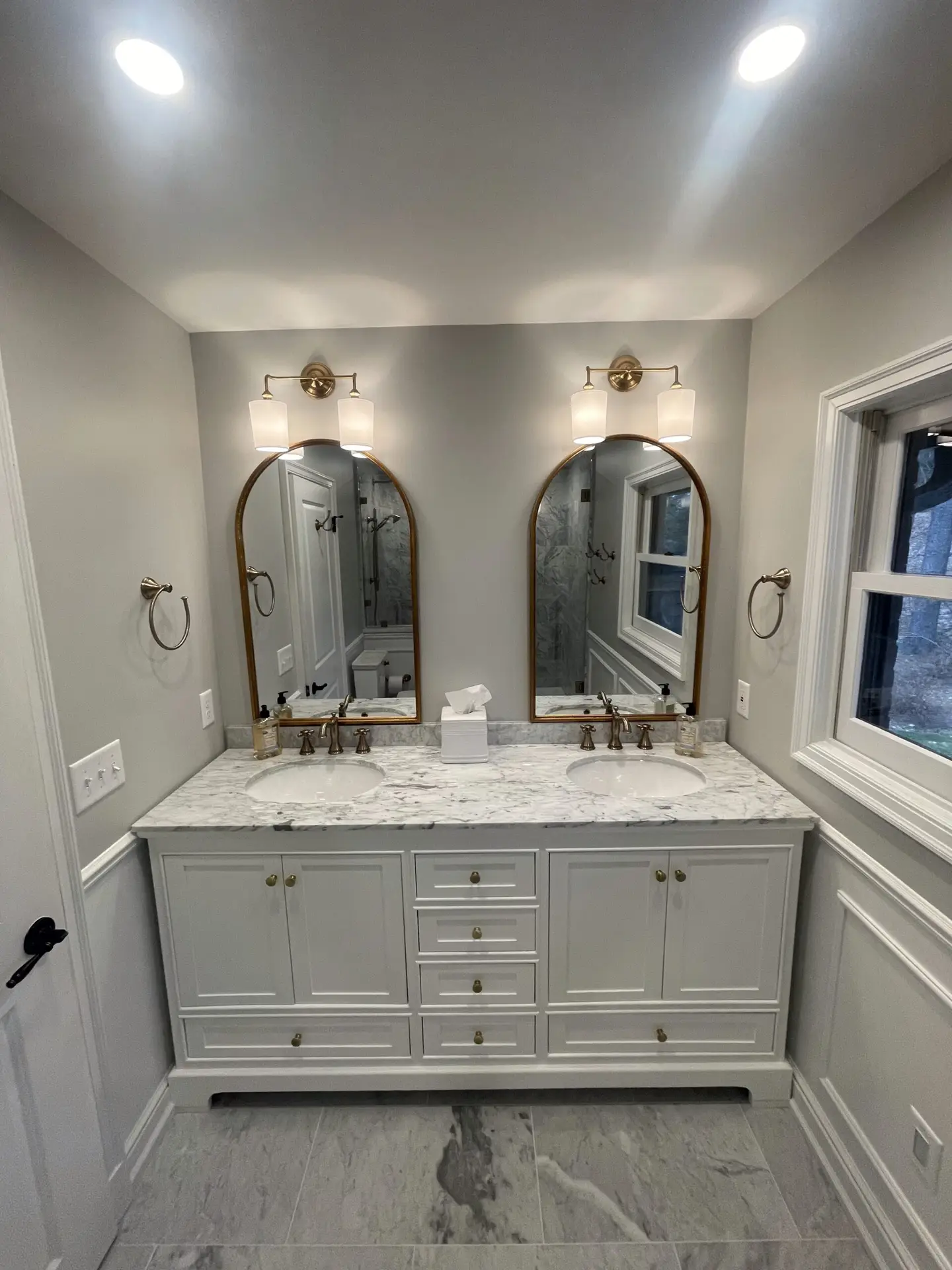 A modern bathroom with a double-sink vanity, marble countertop, two oval mirrors, brass fixtures, wall-mounted lights above each mirror, and a window on the right side. The room has gray walls and tiled floors.
