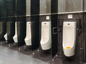 Row of five white urinals mounted on a dark tiled wall in a restroom; the second urinal from the right has black support rails and an orange urinal deodorizer inside.