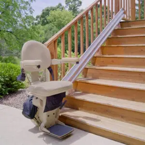 A beige stairlift is installed on the side of an outdoor wooden staircase, surrounded by greenery and trees, providing accessible transportation up the steps.