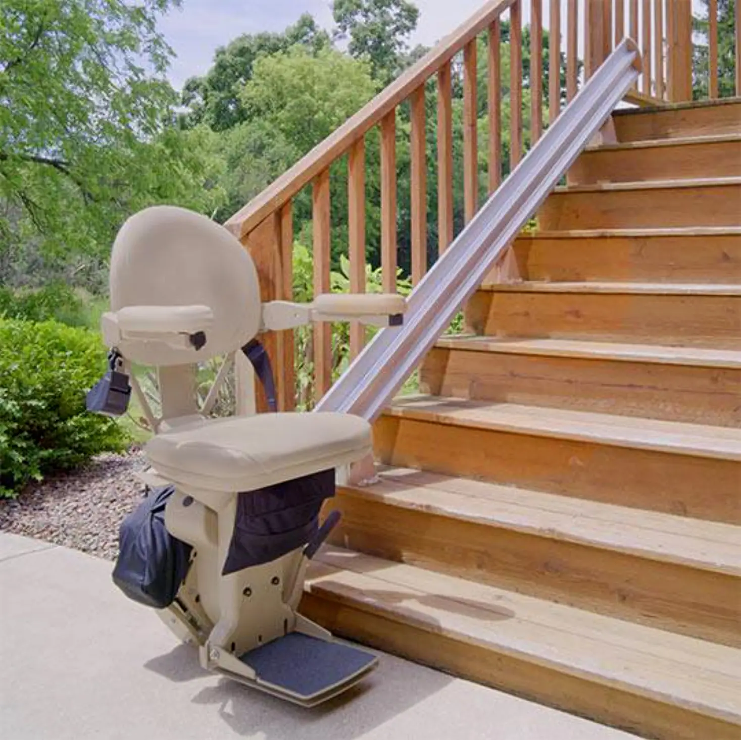 A beige stairlift is installed on the side of an outdoor wooden staircase, surrounded by greenery and trees, providing accessible transportation up the steps.