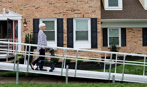 An older adult using a walker moves up a metal wheelchair ramp toward the entrance of a brick house with black shutters and a well-kept yard.