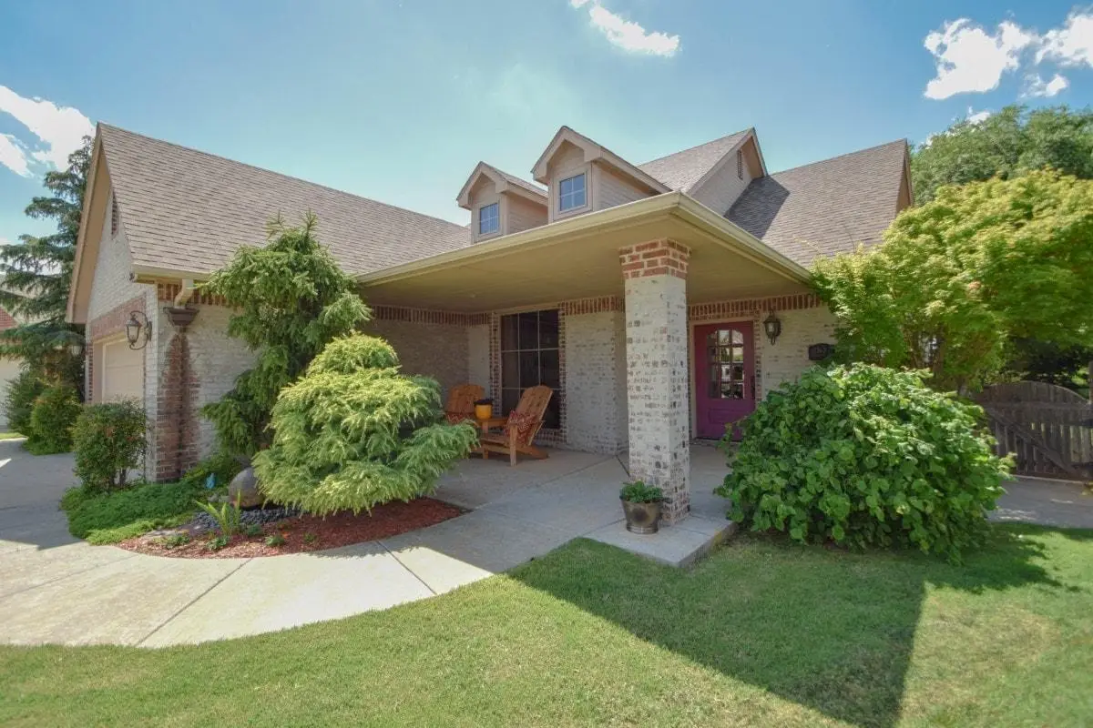 A brick house with a large covered front porch, two wooden chairs, lush green shrubs, and a neatly maintained lawn under a sunny, blue sky.