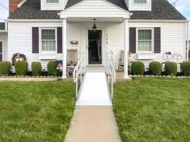 A white house with dark shutters has a front porch and a wheelchair-accessible ramp leading up to the entrance, flanked by neatly trimmed bushes and a well-maintained green lawn.