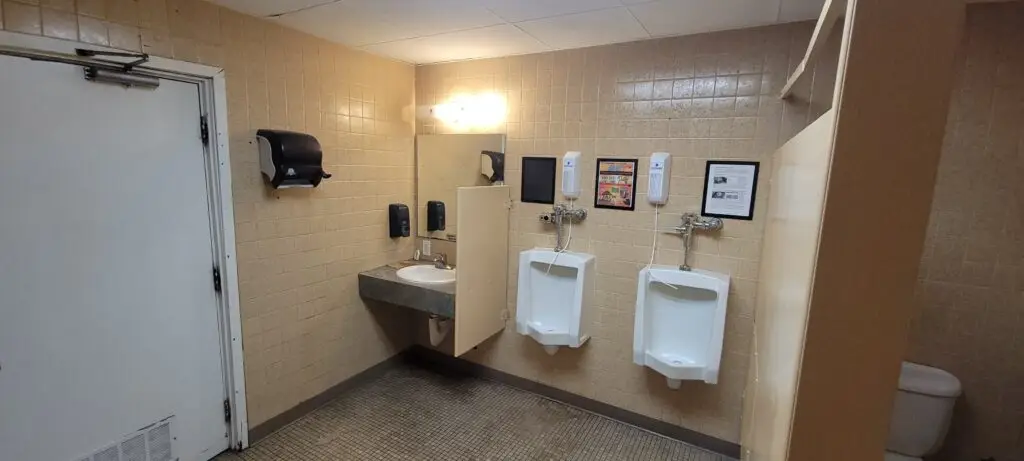 A public restroom with tan tiled walls, featuring a sink with a mirror, soap and paper towel dispensers, two white urinals, framed information on the wall, and a partially visible toilet stall.