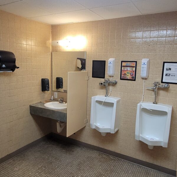 A public restroom with tan tiled walls, featuring a sink with a mirror, soap and paper towel dispensers, two white urinals, framed information on the wall, and a partially visible toilet stall.
