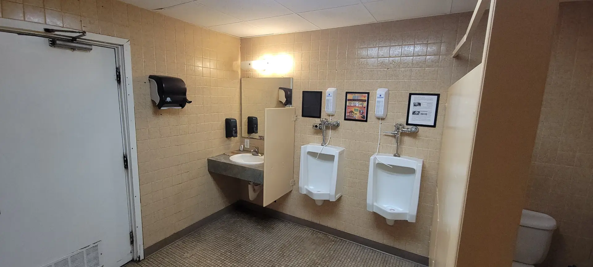 A public restroom with tan tiled walls, featuring a sink with a mirror, soap and paper towel dispensers, two white urinals, framed information on the wall, and a partially visible toilet stall.