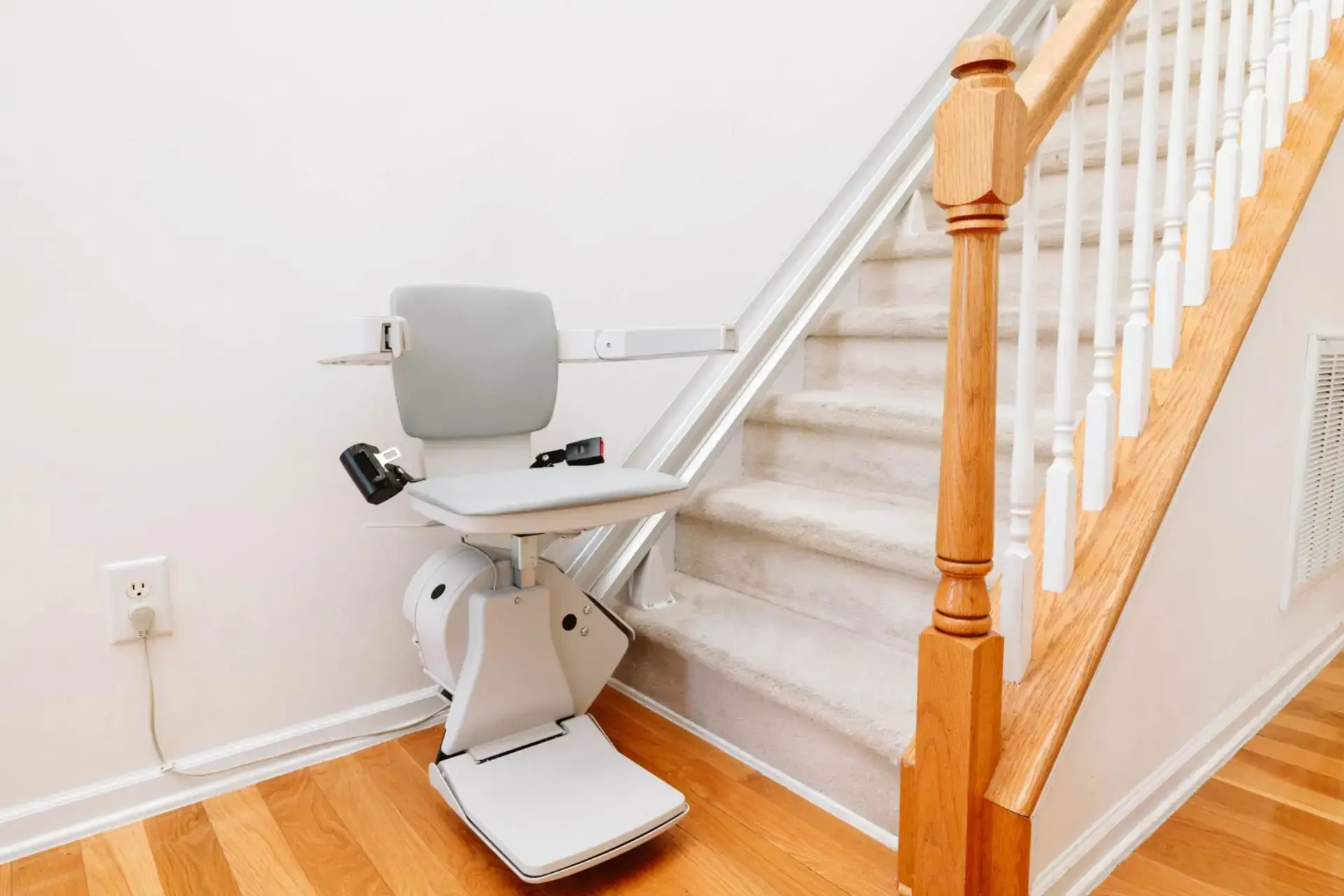 A motorized stair lift with a gray seat and armrests is installed alongside a carpeted staircase with a wooden handrail in a home with hardwood floors and white walls.