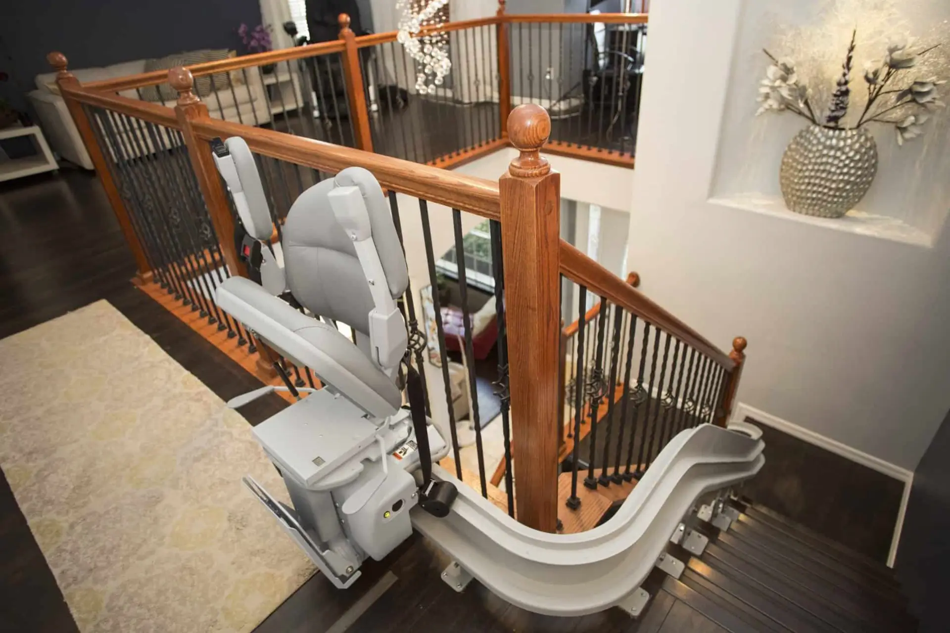 A stairlift with a gray seat and backrest is installed along a curved staircase with dark wooden steps and railings. The stairlift track runs alongside the staircase, designed for safe movement between floors.