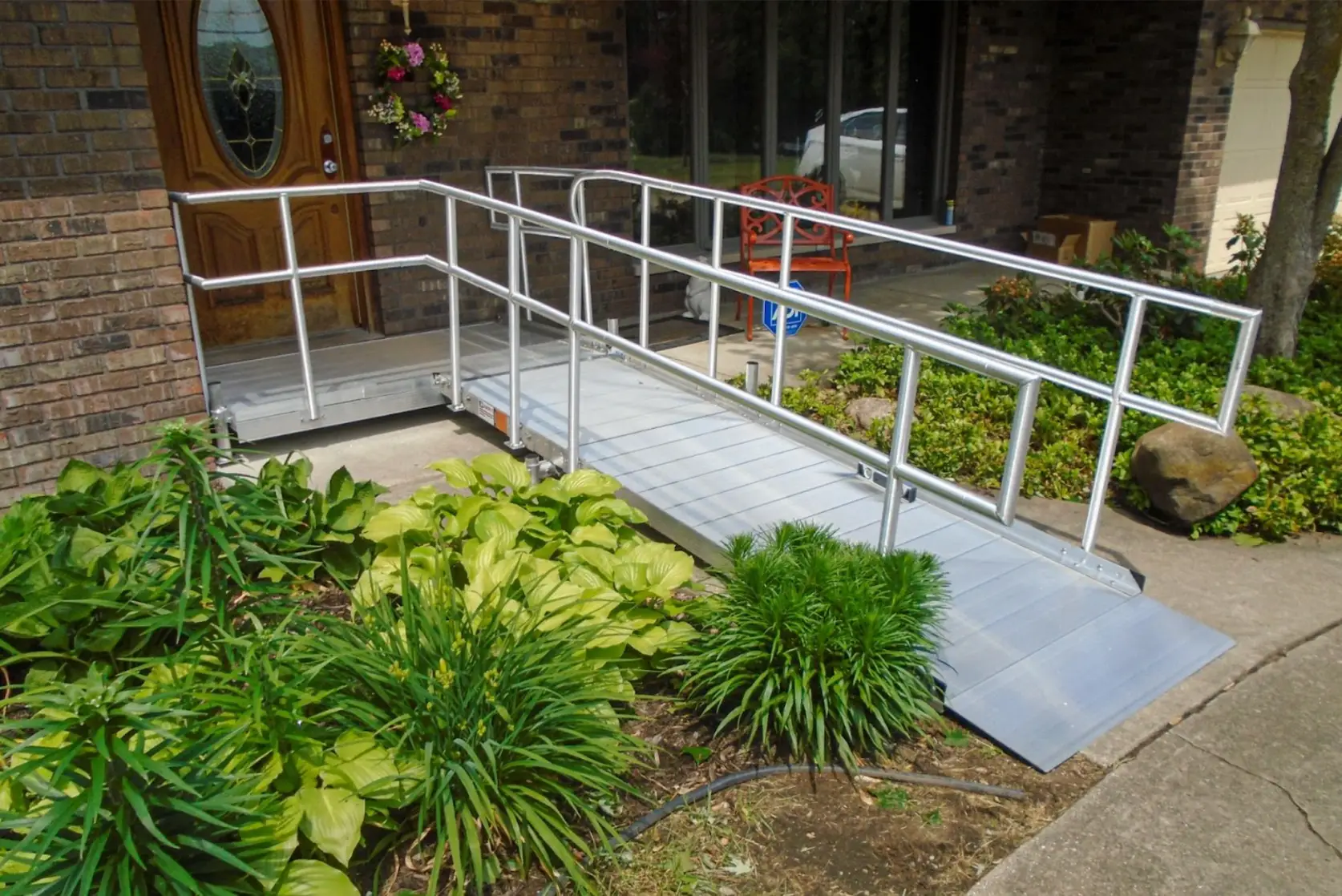 A silver metal wheelchair ramp with handrails leads to the front door of a brick house, surrounded by green plants and shrubs, with a bench and hanging flower basket nearby.