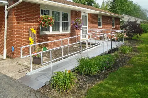 A metal wheelchair ramp with railings leads up to the entrance of a brick house with hanging flower baskets and a small garden along the side.