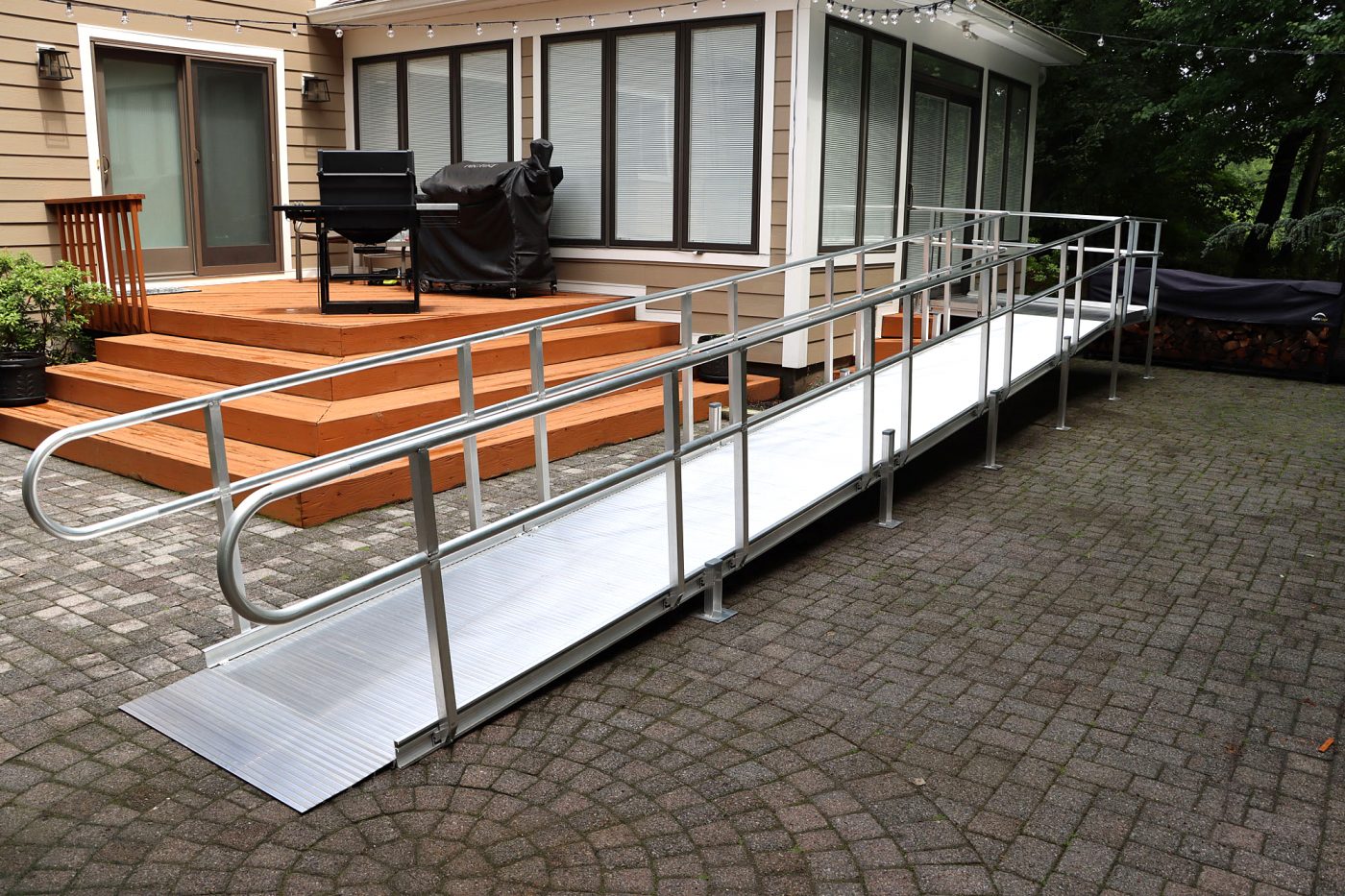 A metal wheelchair ramp with railings leads from a brick patio up to a wooden deck attached to a house with large glass windows.