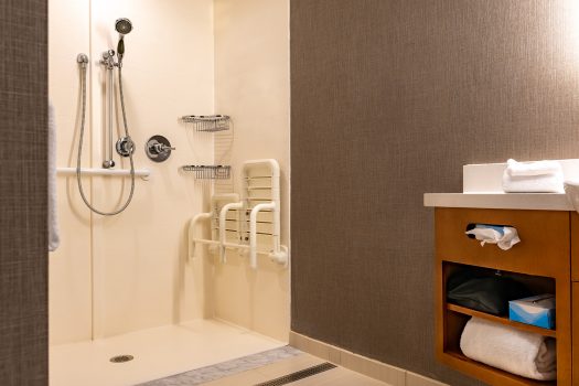 Accessible hotel bathroom with a walk-in shower, grab bars, a fold-down shower seat, handheld showerhead, and open shelving with towels and toiletries next to the sink. Accessible hotel bathroom with a walk-in shower, grab bars, a fold-down shower seat, handheld showerhead, and open shelving with towels and toiletries next to the sink.